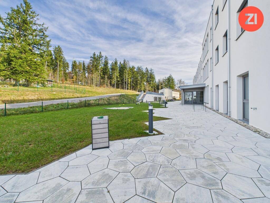 Weitläufiger Außenbereich mit modernem Gebäude, gepflasterten Wegen und angrenzender Naturlandschaft mit Bäumen.