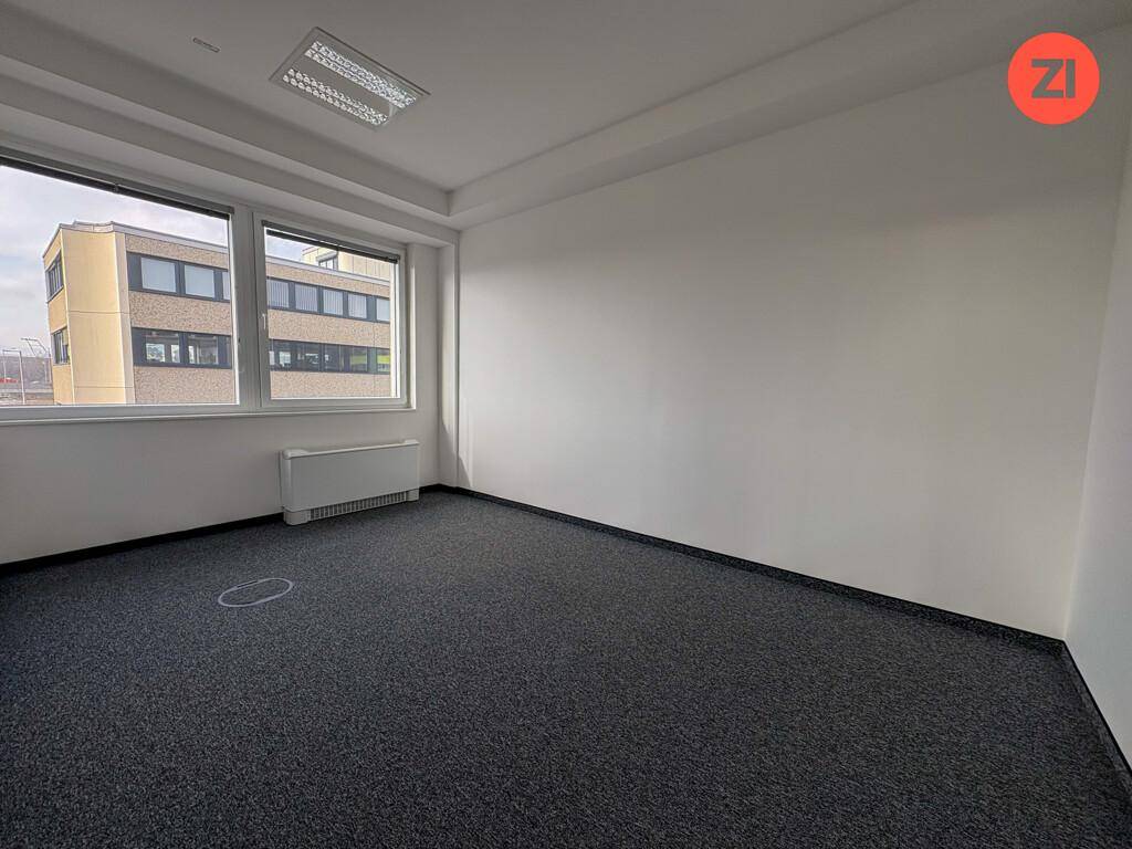 Helles Büro mit Teppichboden und einem Fenster, das einen Blick auf ein angrenzendes Gebäude bietet.