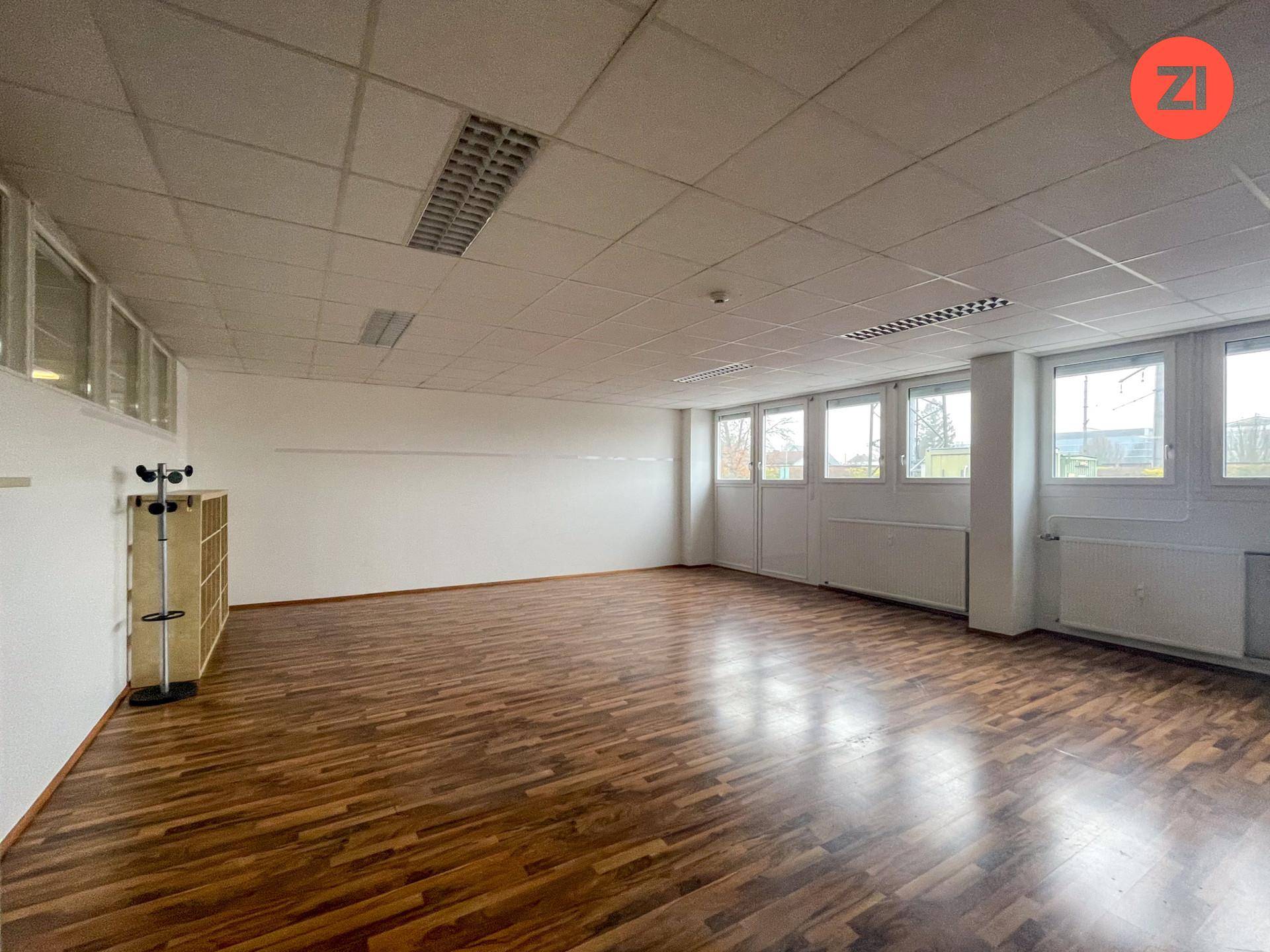 Helles Büro mit Holzoptik-Boden, vielen Fenstern und einem einfachen Regal an der Wand.