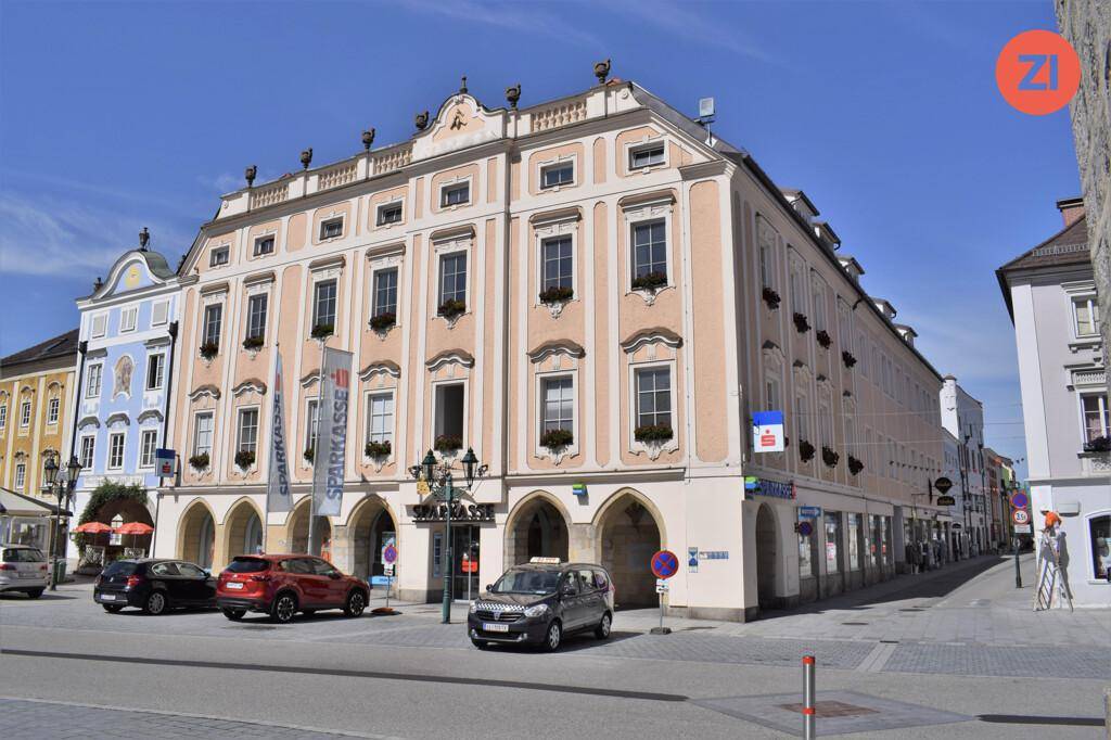 Historisches Gebäude mit eleganter Fassade und Arkaden im Erdgeschoss, an einem sonnigen Marktplatz gelegen.