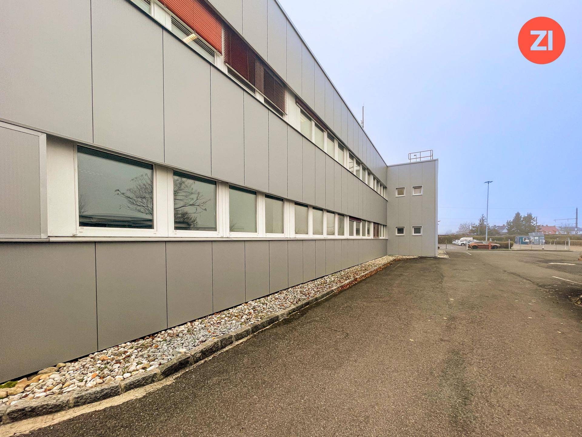 Moderne Gewerbefassade mit langen Fensterreihen, grauen Paneelen und einem Kiesstreifen am Gebäude.