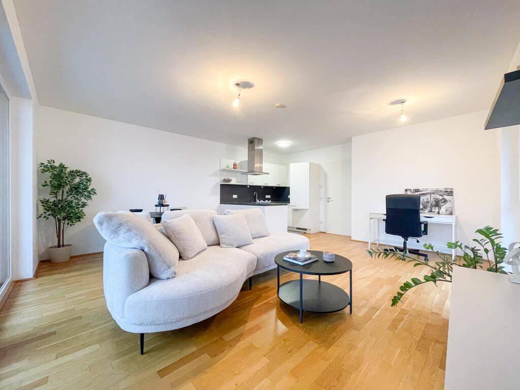 Lichtdurchfluteter Wohnbereich mit offener Küche, modernem Sofa und einem Arbeitsplatz am Fenster.