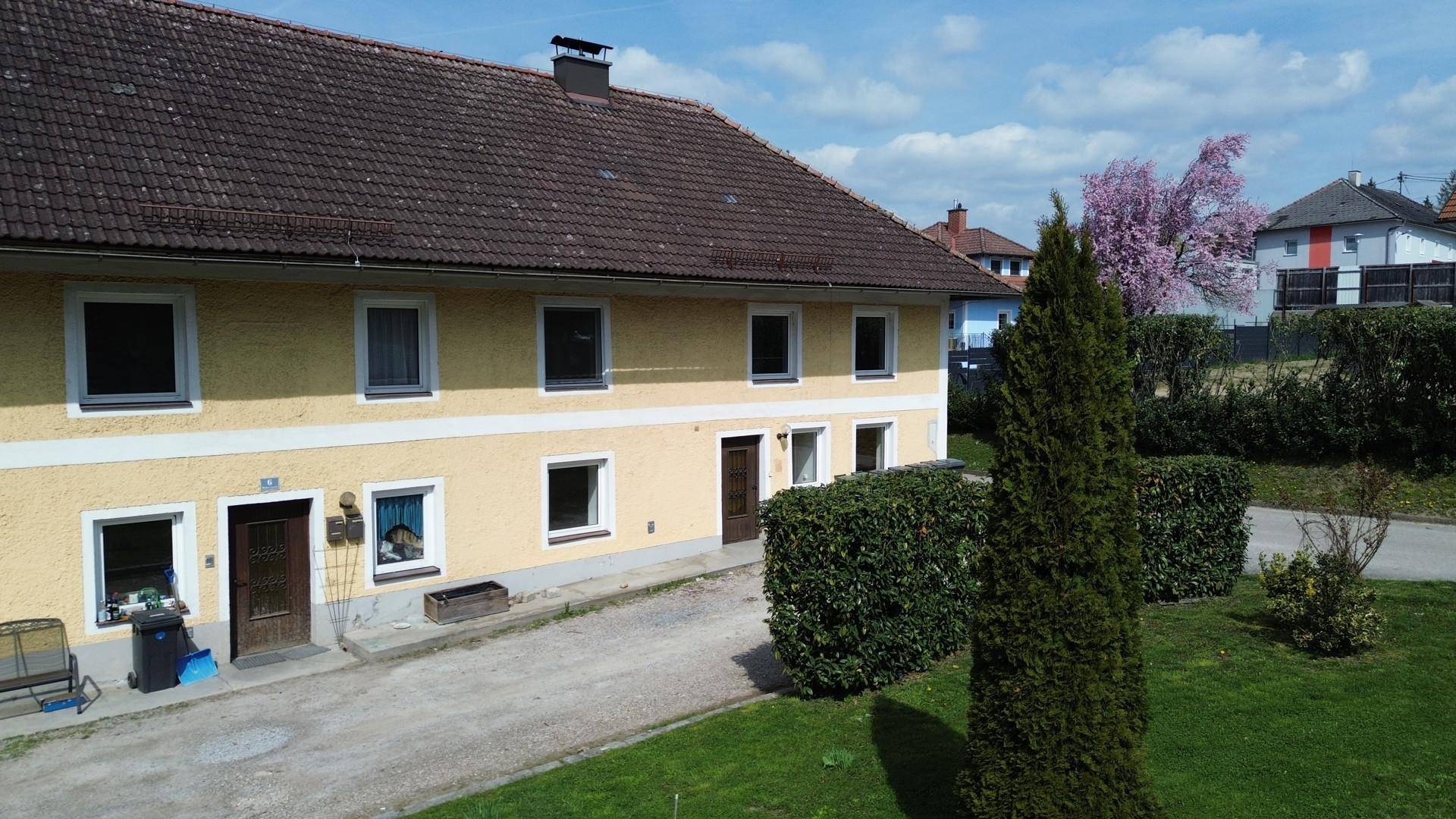 Gelbes Gebäude mit braunem Ziegeldach, umgeben von einer grünen Wiese und Hecken unter blauem Himmel.