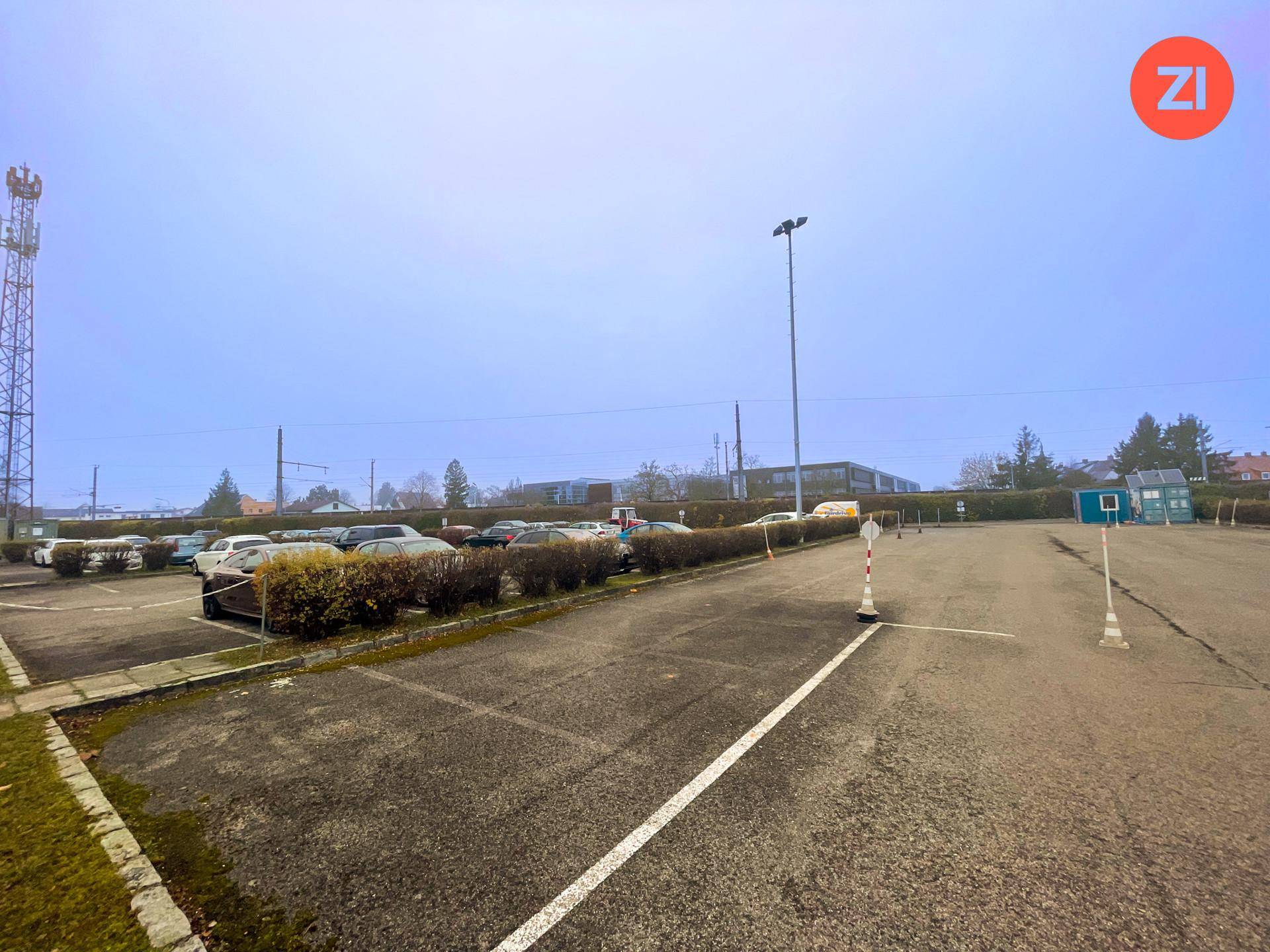 Großer Außenparkplatz mit vielen Fahrzeugen, einem Mobilfunkmast und einer weiten Himmelsansicht.