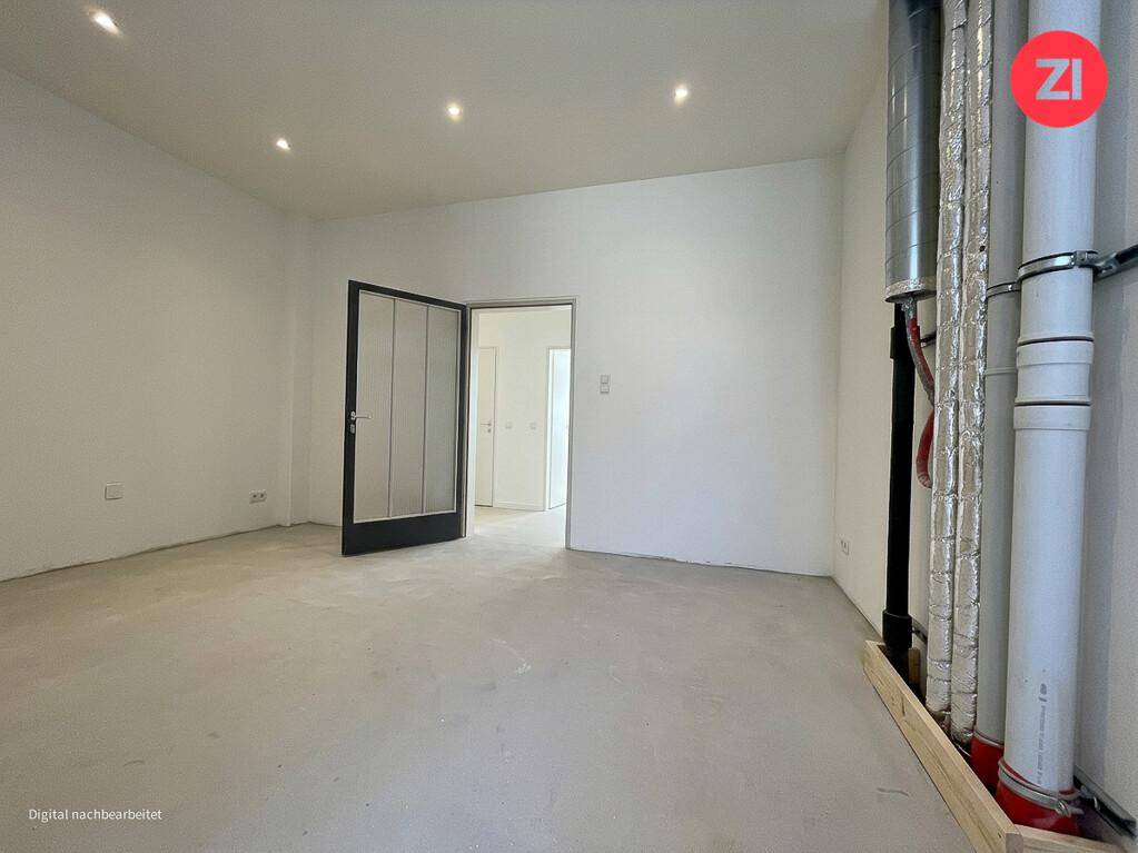 Leerer Raum mit Betonboden, weißen Wänden und sichtbaren Versorgungsleitungen, bereit für den Ausbau.