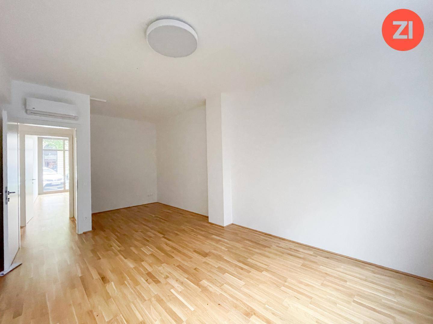 Leerer Raum mit Holzboden, weißen Wänden und einer Klimaanlage über dem Türrahmen.