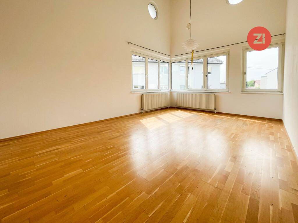 Geräumiger, heller Raum mit Holzparkett, hohen Decken und vielen Fenstern, die viel Tageslicht hereinlassen.