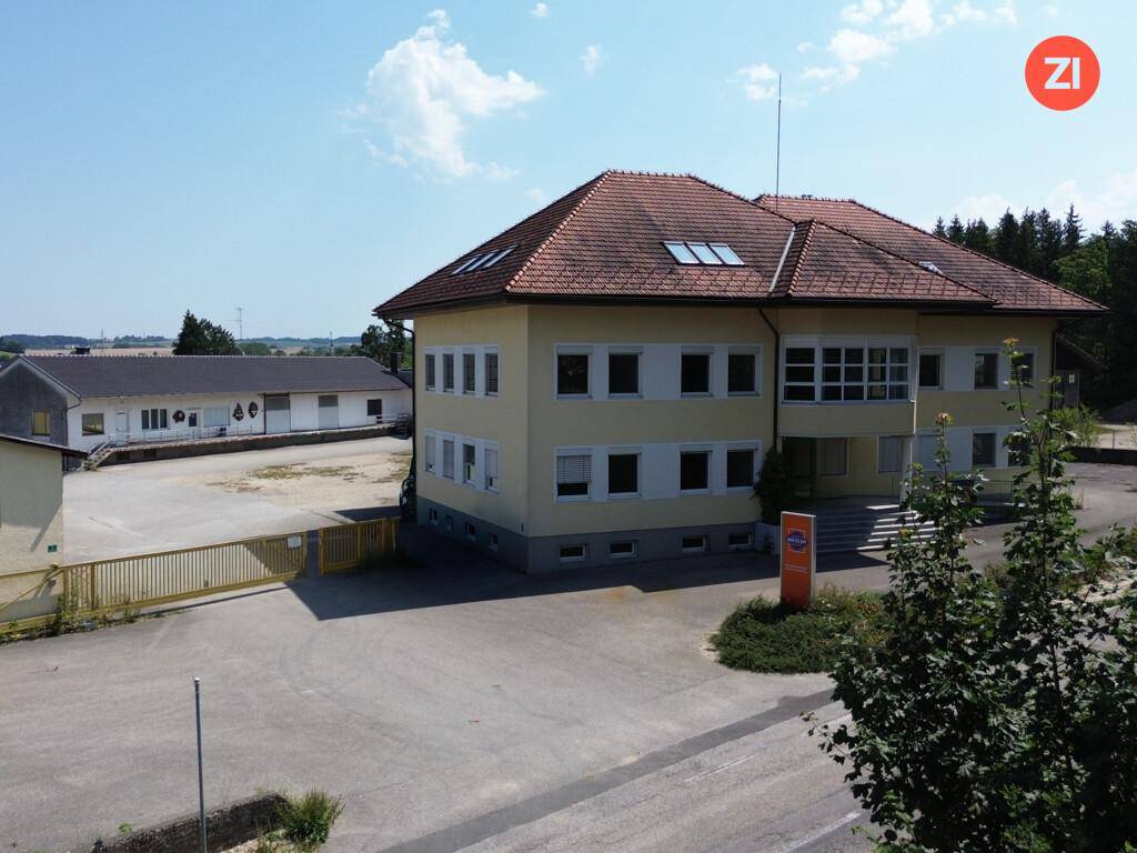 Hauptgebäude mit gelber Fassade und rotem Ziegeldach, umgeben von einem großen, unbefestigten Parkplatzbereich.