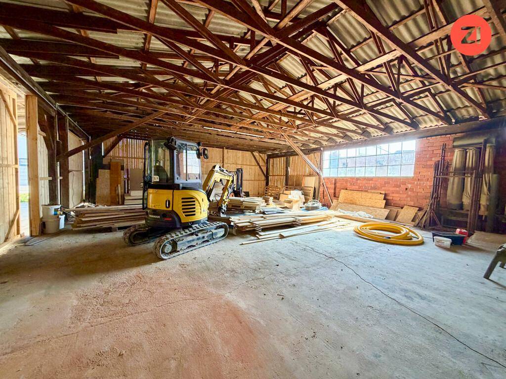 Großer Lagerraum mit sichtbarer Holzkonstruktion des Daches und einem kleinen Bagger auf dem Betonboden.
