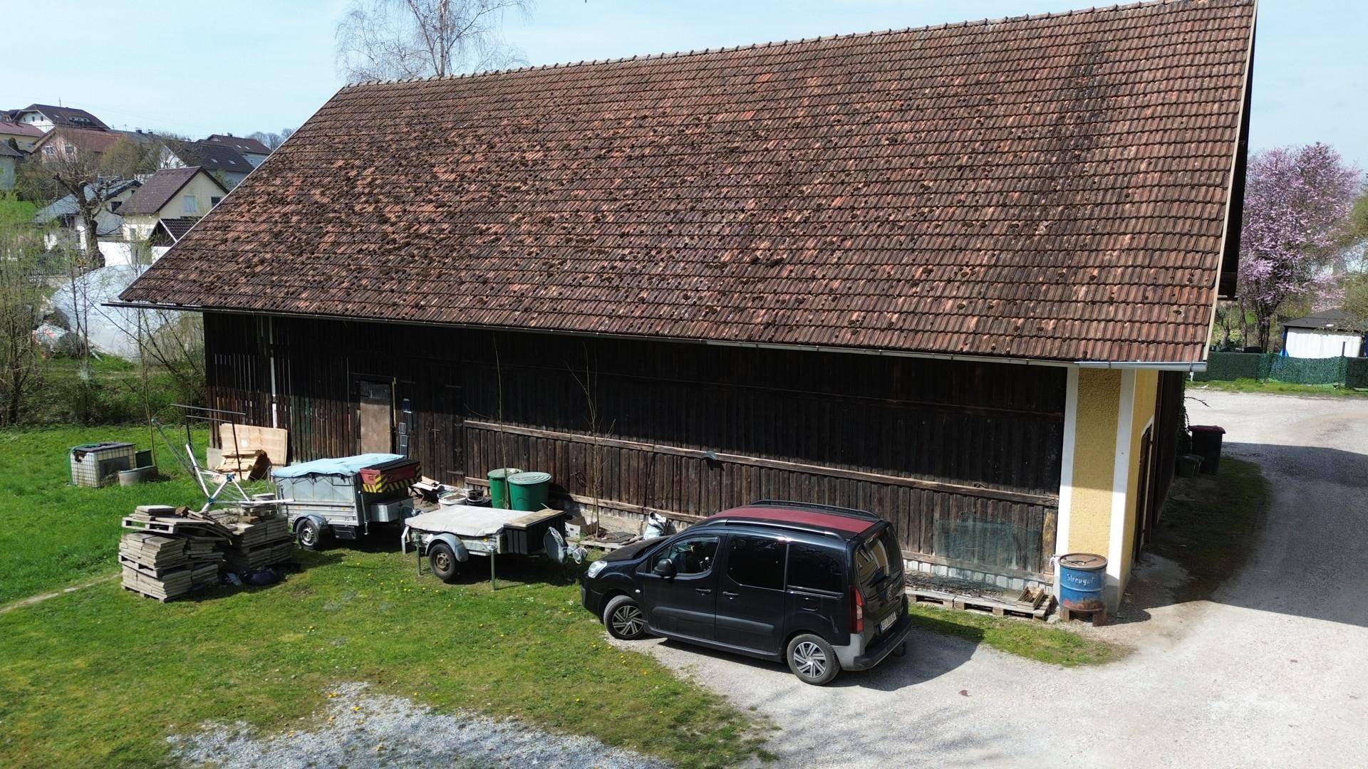 Großes Scheunengebäude aus dunklem Holz mit Ziegeldach, umgeben von einer Wiese und Fahrzeugen.