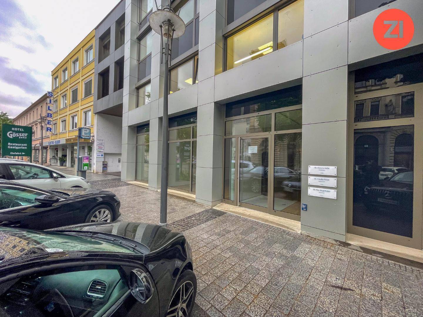 Außenansicht eines modernen Geschäftsgebäudes an einer Straße mit parkenden Autos und angrenzenden Gebäuden.
