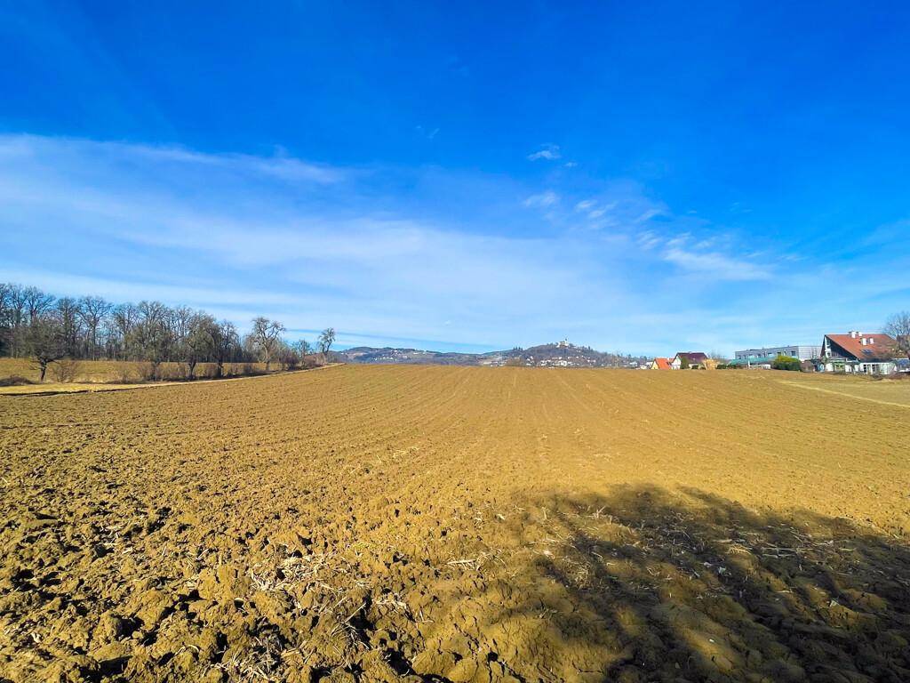 Weitläufiges Ackerland mit Blick auf eine hügelige Landschaft und vereinzelte Häuser unter blauem Himmel.