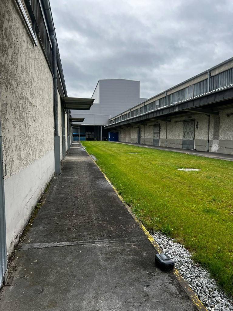 Langer gepflasterter Weg entlang eines Industriegebäudes mit Grünstreifen und weiteren Gebäuden in der Ferne.