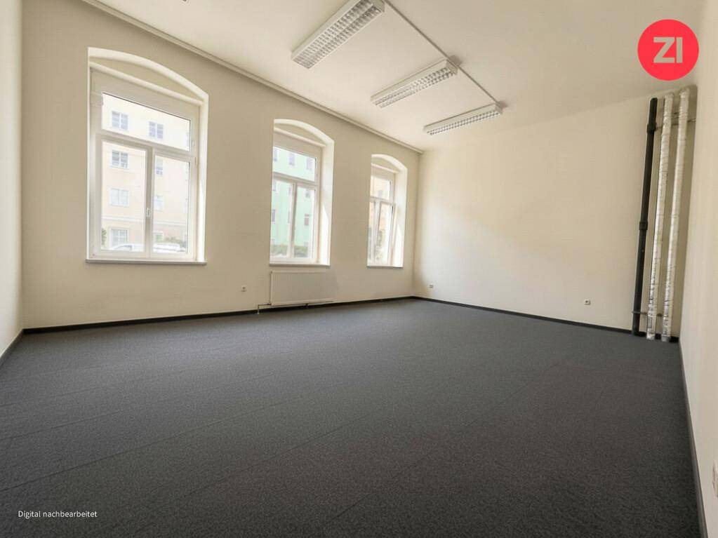 Leerer Büroraum mit dunklem Teppichboden, großen Fenstern und Deckenbeleuchtung. Der Raum ist hell und funktional.