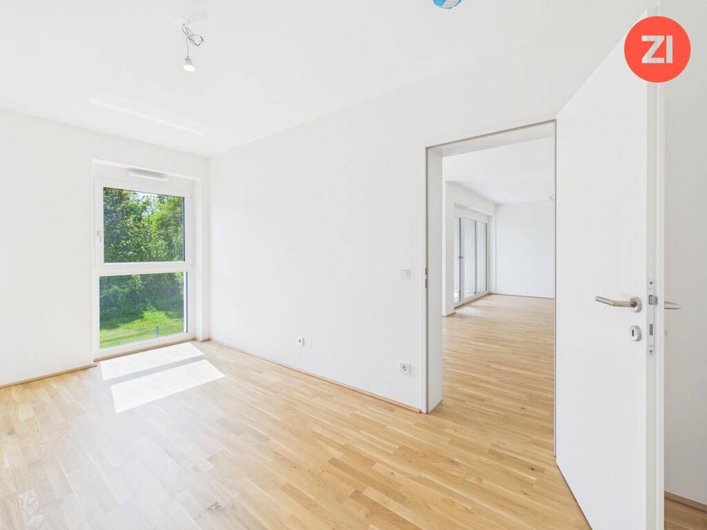 Helles Zimmer mit Holzboden und großem Fenster, das viel Sonnenlicht hereinlässt und einen Grünblick bietet.