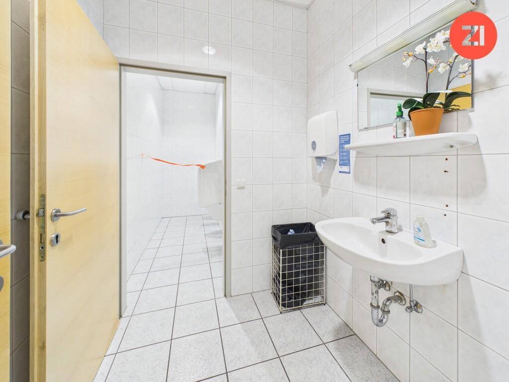 Helles Badezimmer mit weißen Fliesen, Waschbecken, Spiegel und Zugang zu weiteren Toiletten.
