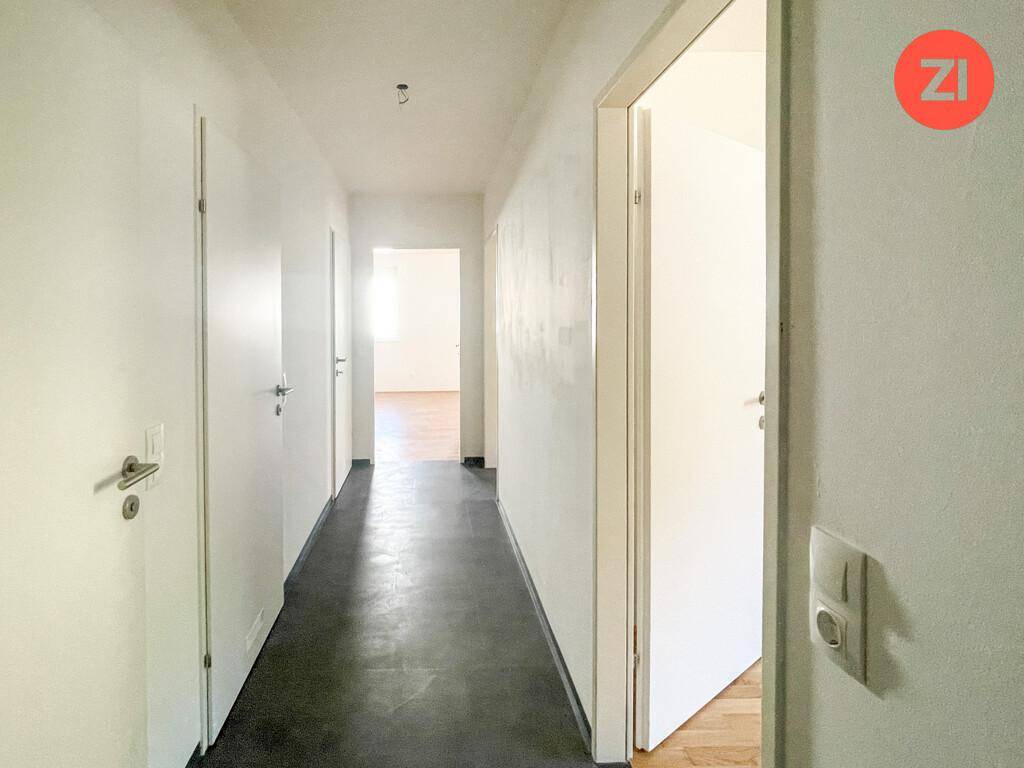 Heller Flur mit weißen Wänden, dunklem Boden und Zugang zu weiteren Zimmern.