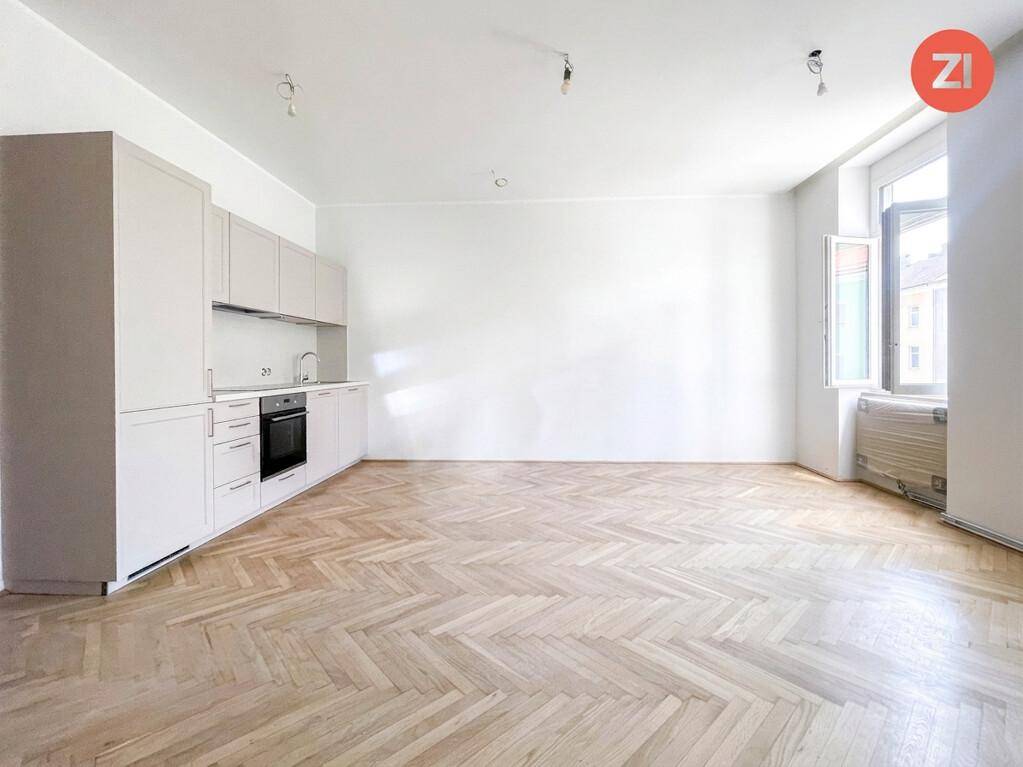 Geräumiger Wohnbereich mit Fischgrätparkett, integrierter Küchenzeile und großem Fenster für viel Tageslicht.