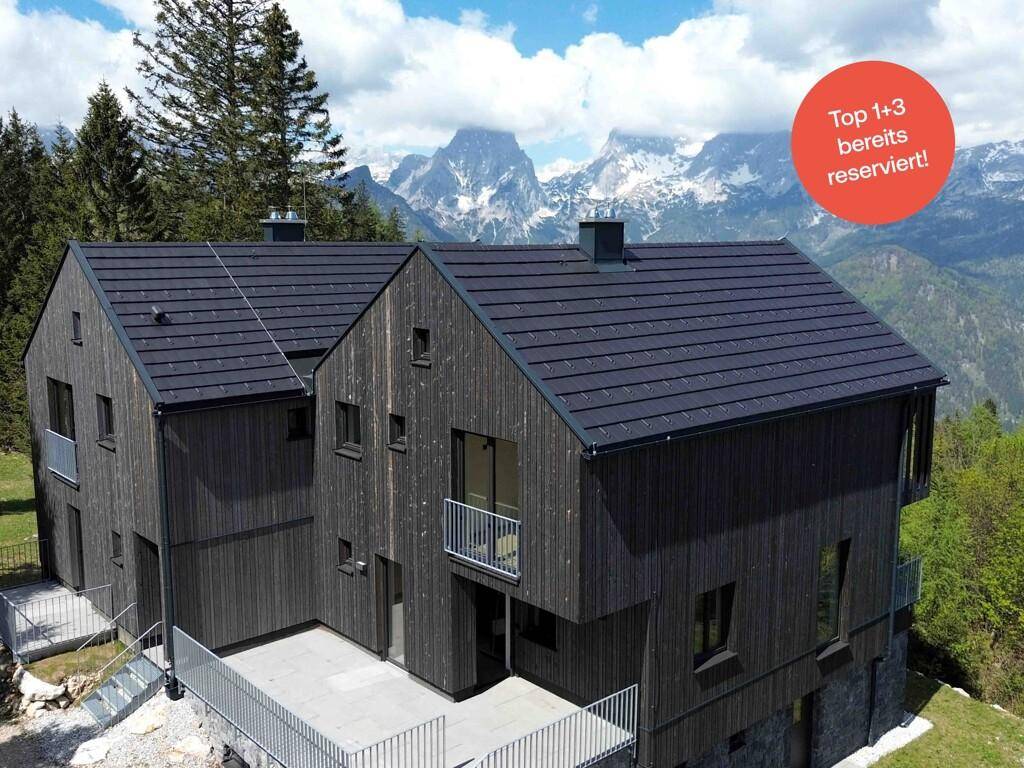 Modernes Haus mit dunkler Holzfassade, Satteldach und großer Terrasse, umgeben von einer beeindruckenden Berglandschaft.