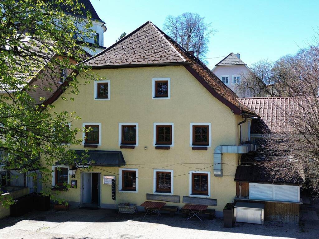 Traditionelles gelbes Gebäude mit braunen Fenstern und einem Außenbereich, umgeben von Bäumen und Grün.