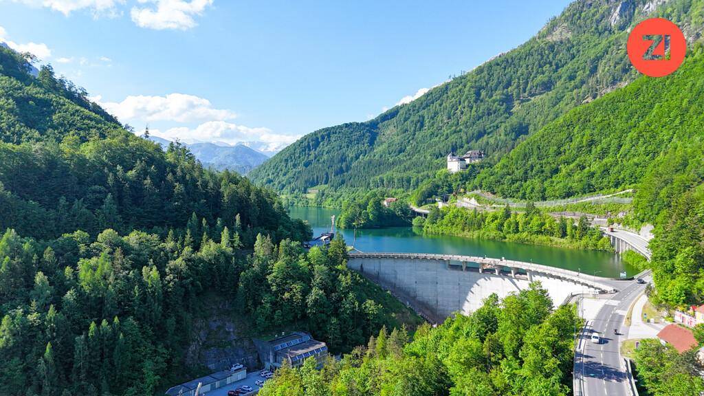 Beeindruckende Luftaufnahme eines Stausees in einem grünen Tal, umgeben von bewaldeten Bergen.
