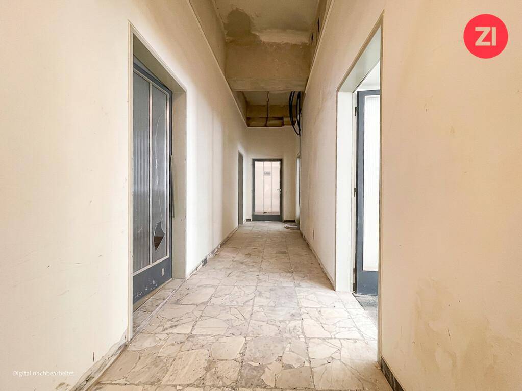 Langer Flur mit mehreren Türen, Fliesenboden und sichtbaren Deckeninstallationen, möglicherweise in Renovierung. Der Zustand ist original.