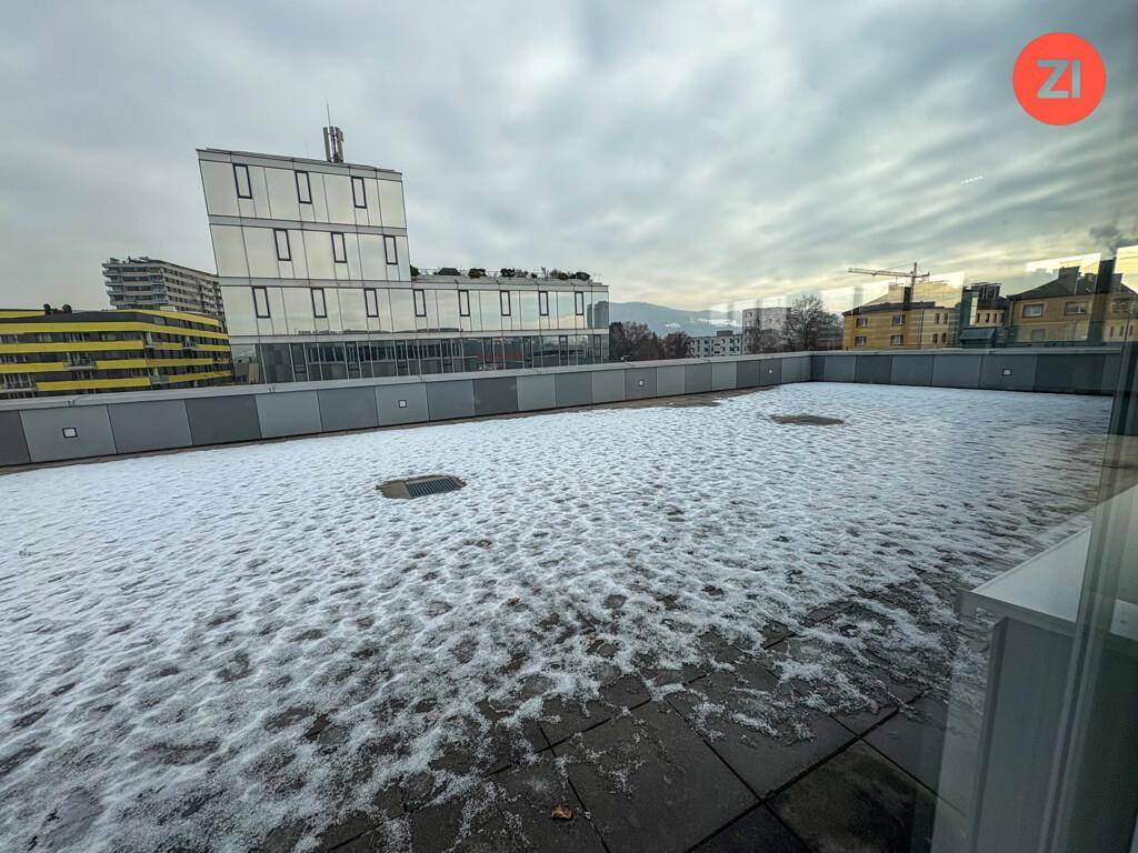 Dachterrasse mit Schnee bedeckt und Blick auf umliegende Gebäude und Berge im Hintergrund.