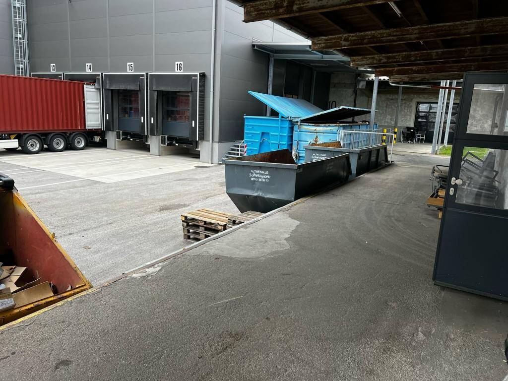 Laderampenbereich eines Industriegebäudes mit mehreren Toren und einem LKW, daneben große Abfallcontainer.