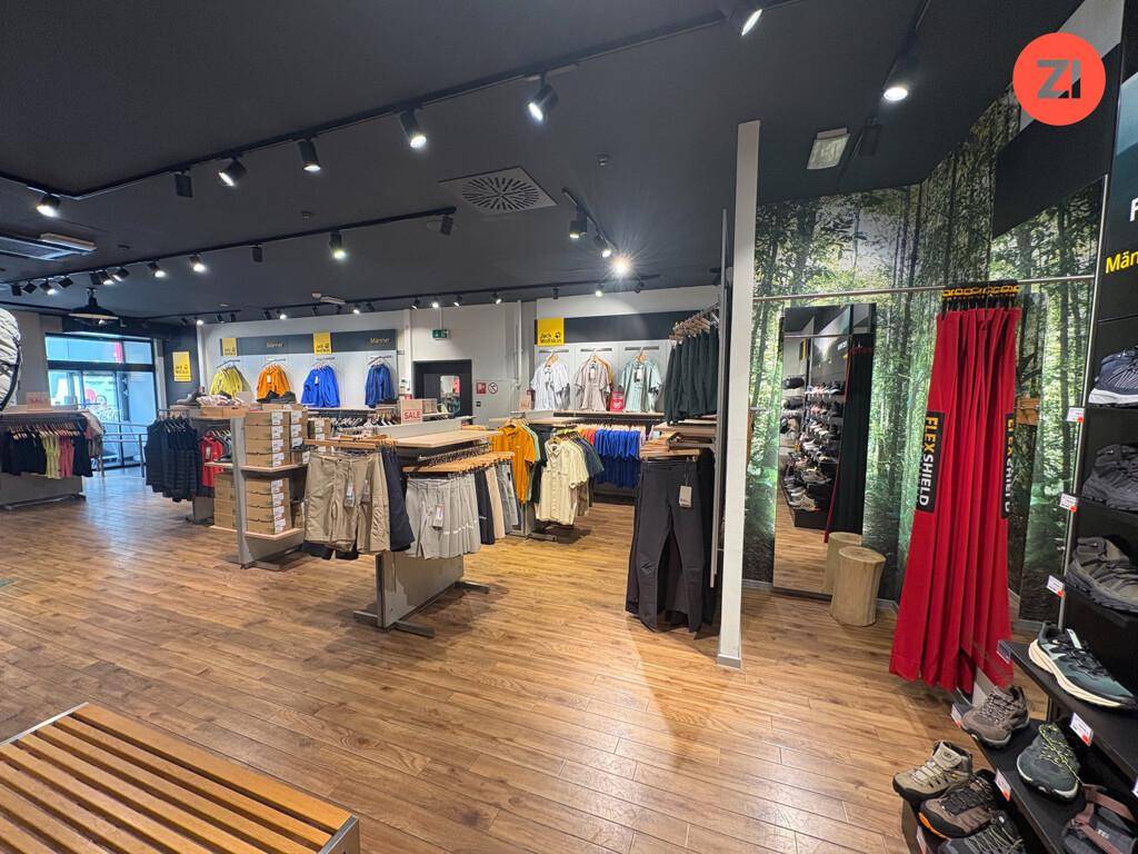 Geräumiger Verkaufsraum eines Bekleidungsgeschäfts mit Holzfußboden, modernen Regalen und Waldmotiv-Tapete.