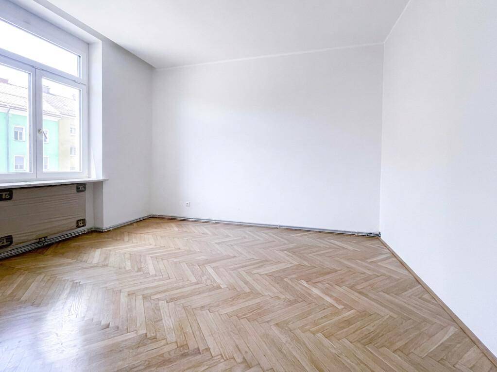 Leerer Raum mit klassischem Fischgrätparkett und einem Fenster, das für eine angenehme Helligkeit sorgt.