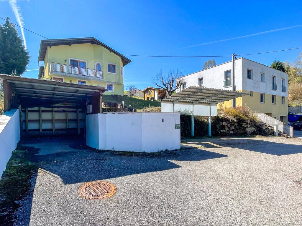 Gelbes Haus mit Parkplätzen und Carport in Hanglage, umgeben von weiterer Bebauung.