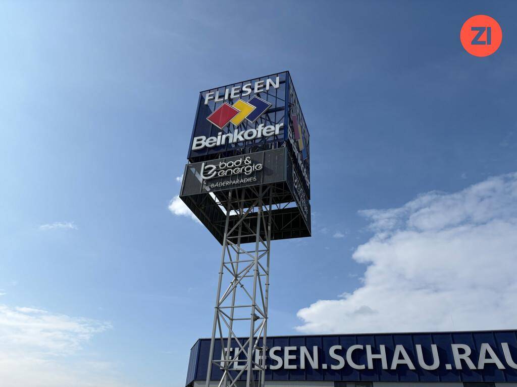 Hoher Werbepylon mit Firmenlogos vor einem blauen Himmel und Geschäftsgebäude.