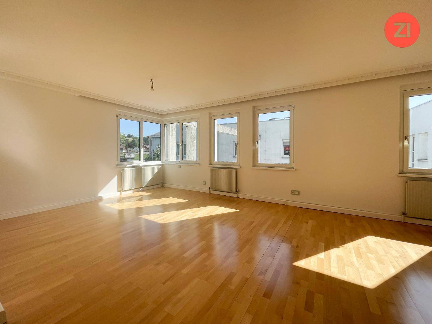 Heller, leerer Raum mit Parkettboden und vielen Fenstern, die viel Tageslicht hereinlassen.