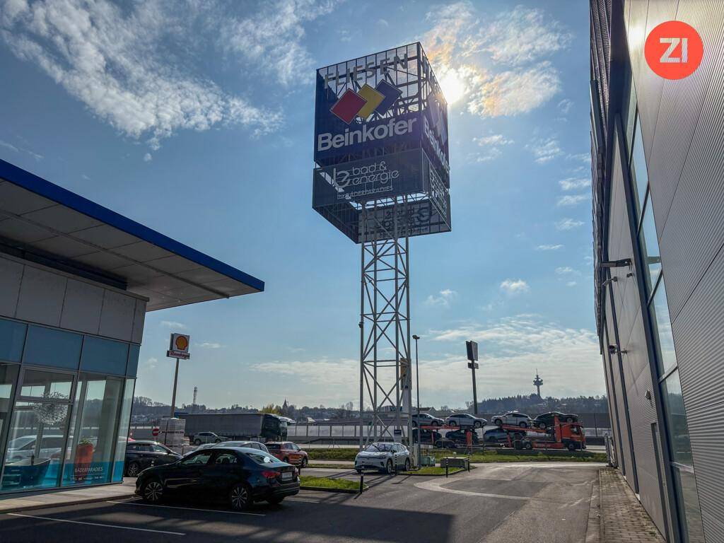 Hoher Werbepylon mit Firmenlogos und umliegendem Parkplatz unter sonnigem Himmel.
