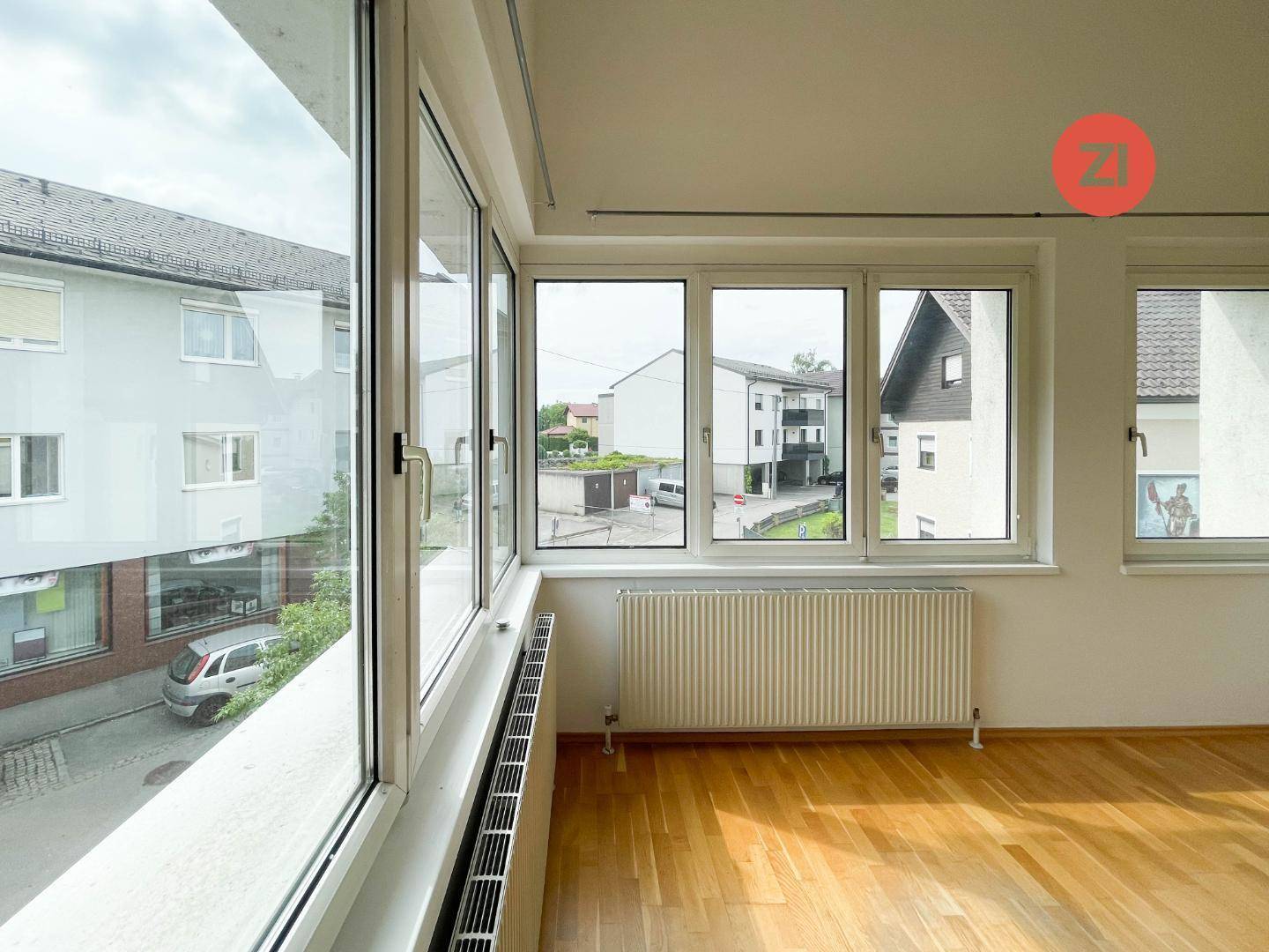 Blick aus dem Wohnzimmer auf die Straße und umliegende Gebäude, mit Holzboden und Heizkörper.