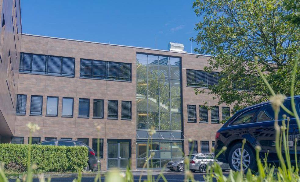 Modernes Bürogebäude mit brauner Fassade, großen Fenstern und einem prominenten Glas-Eingangsbereich, umgeben von Grünflächen und Parkplätzen.