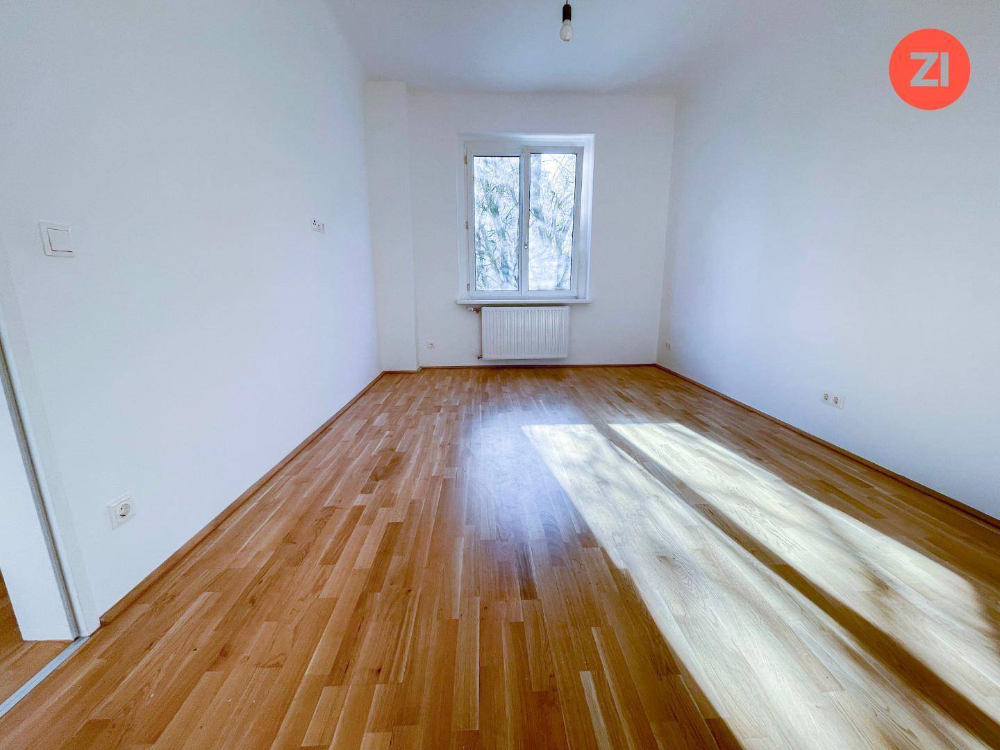 Leeres Zimmer mit hellem Parkettboden und einem großen Fenster, das den Raum gut belichtet.