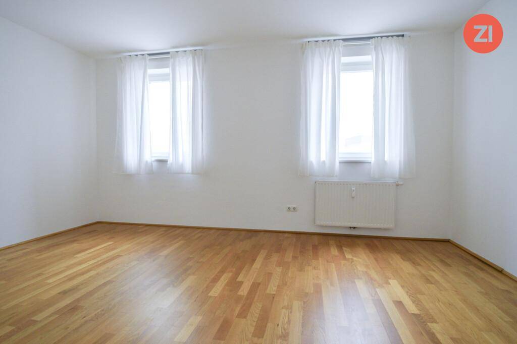 Helles Zimmer mit zwei Fenstern, Heizkörper und klassischem Parkettboden.
