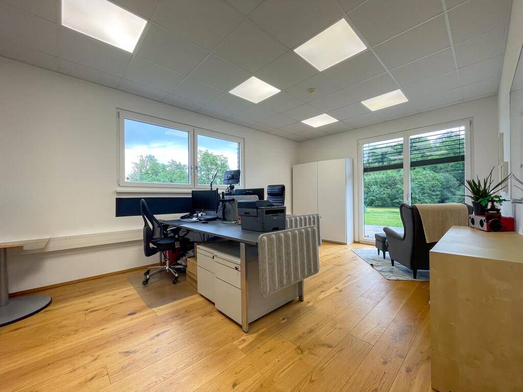 Helles Büro mit Holzboden, modernem Schreibtisch und großen Fenstern mit Blick ins Grüne.
