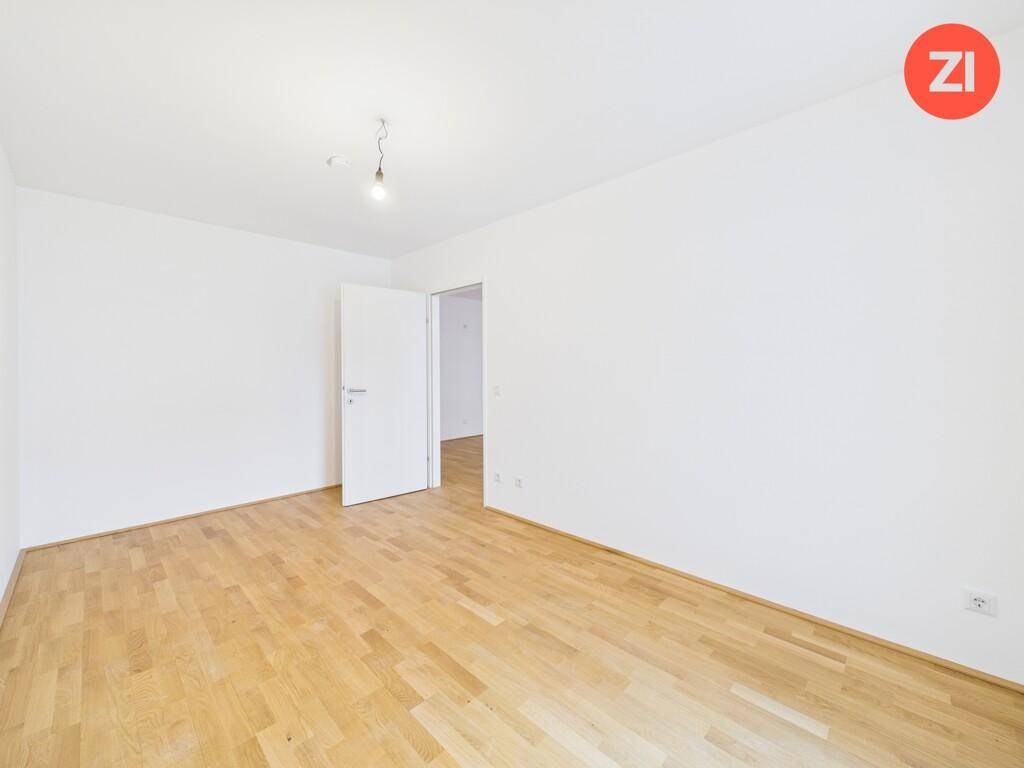 Leerer Raum mit hellem Parkettboden, weißen Wänden und einer offenen Tür zu einem weiteren Bereich.