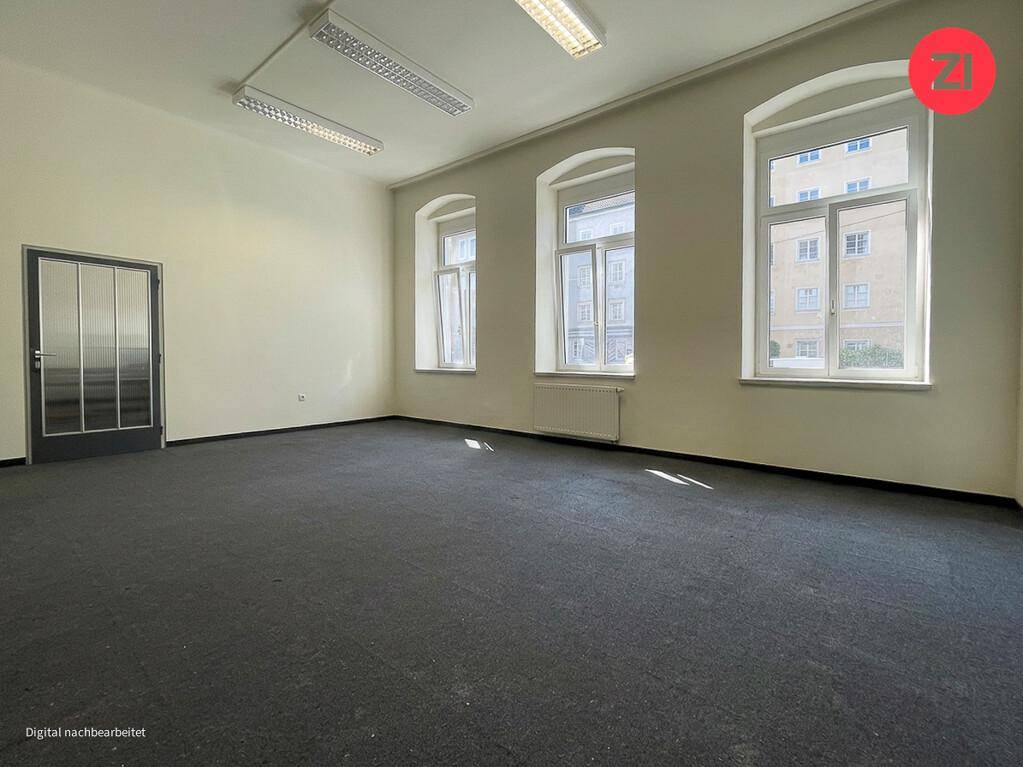 Geräumiger Büroraum mit Teppichboden, großen Fenstern und einer modernen Glastür.