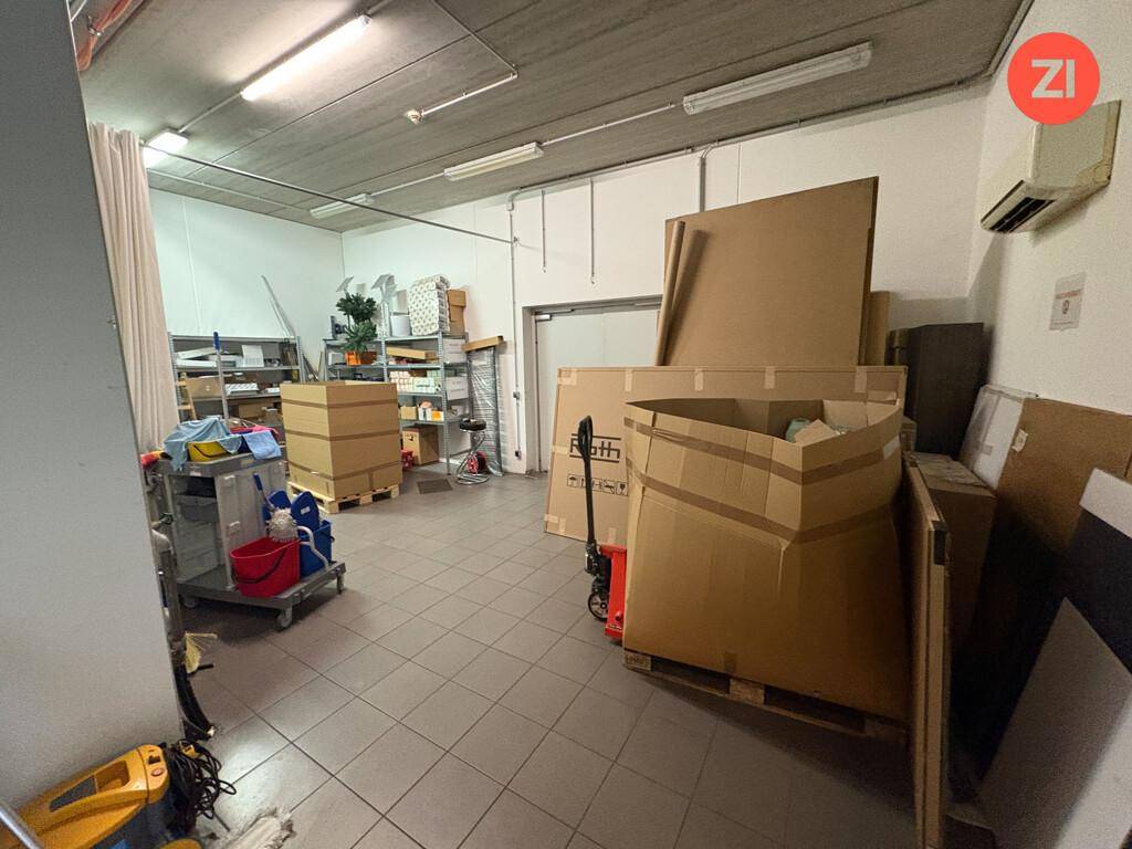 Lagerraum mit Kartons, Regalen und verschiedenen Materialien, gekennzeichnet durch einen Fliesenboden und einfache Beleuchtung.
