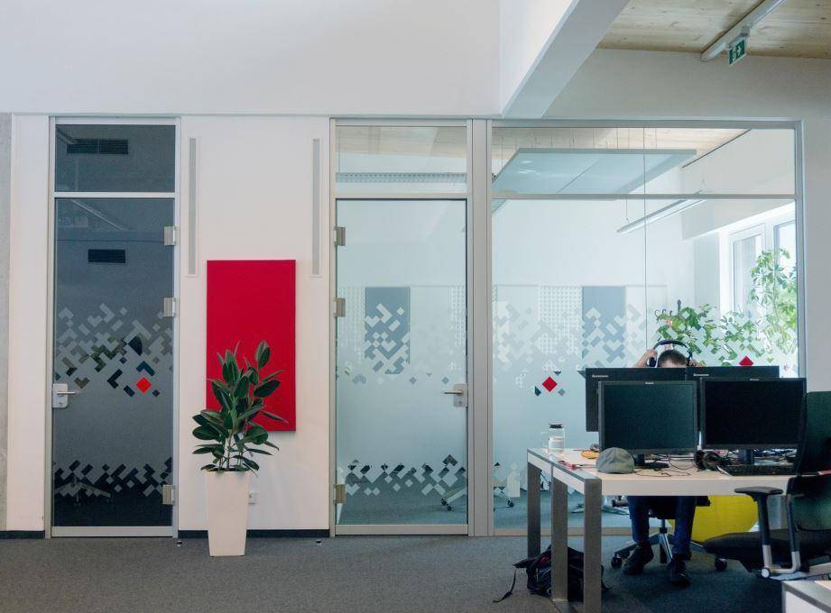 Moderner Bürobereich mit Glaswänden und Türen, die für Transparenz sorgen, sowie einem roten Akzent an der Wand und grünen Pflanzen.