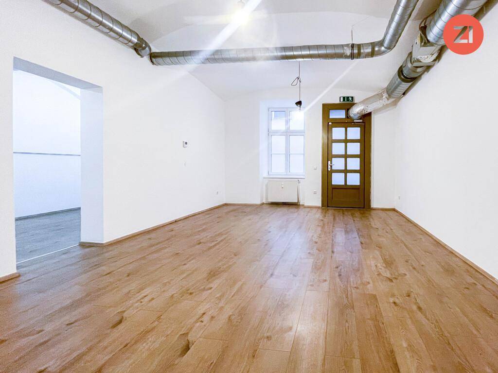 Leerer Raum mit Holzboden, weißen Wänden, sichtbaren Lüftungsrohren und einem Fenster.