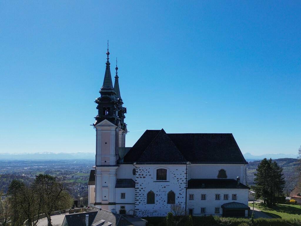 Luftaufnahme einer historischen Kirche mit zwei Türmen, eingebettet in eine grüne Hügellandschaft.