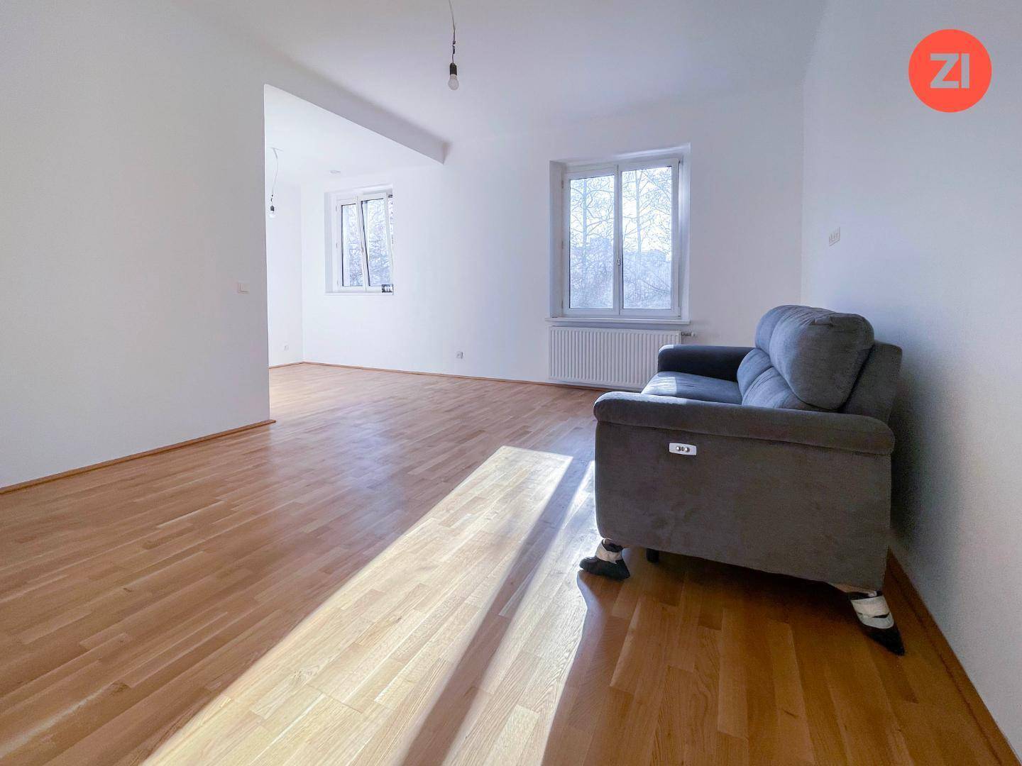 Geräumiges Wohnzimmer mit Holzboden und zwei Fenstern, die den Raum mit natürlichem Licht füllen.