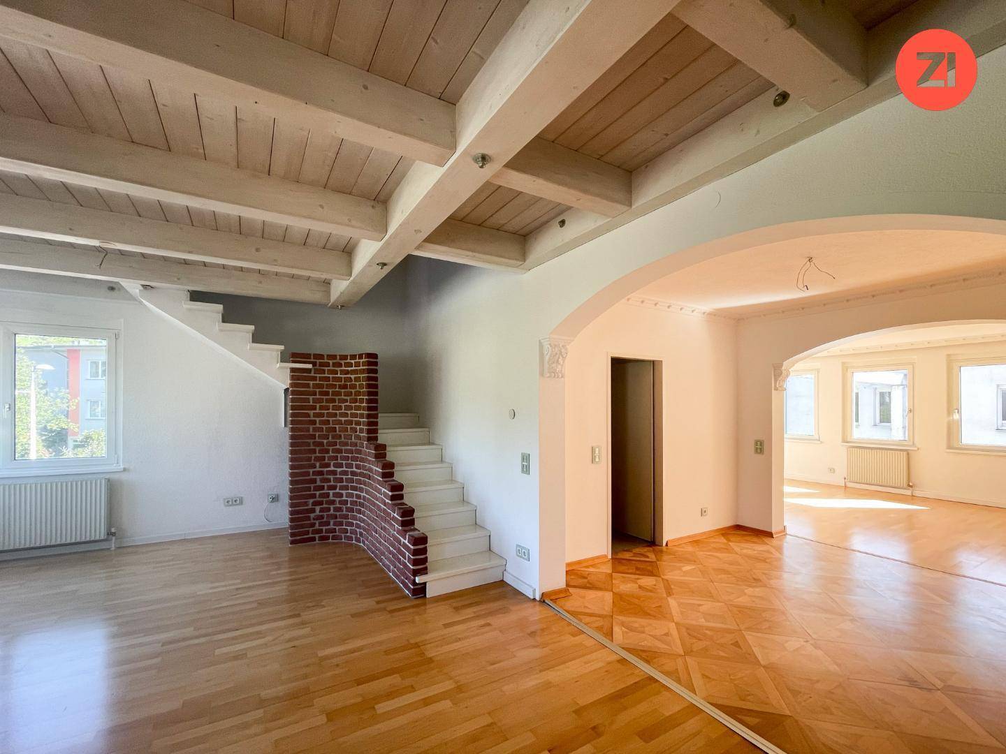 Geräumiges Wohnzimmer mit Holzbalkendecke, Parkettboden und einem eleganten Bogen zum angrenzenden Bereich.