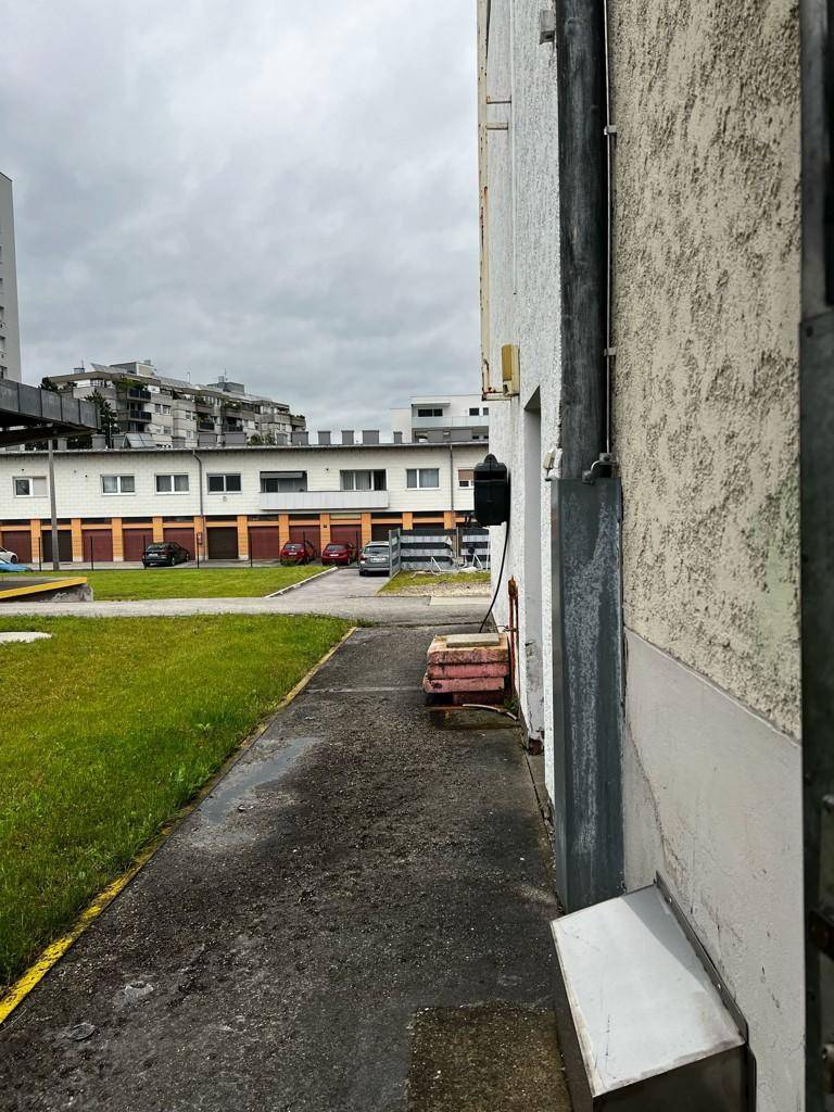 Seitenansicht eines Gebäudes mit schmalem Gehweg und Grünstreifen, im Hintergrund weitere Gebäude und Wohnbebauung.