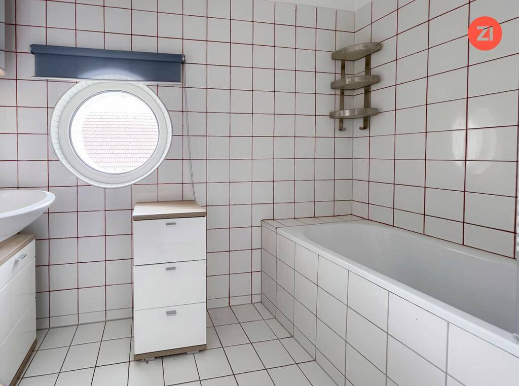 Badezimmer mit weißen Fliesen, Badewanne, Waschbecken und einem markanten runden Fenster.