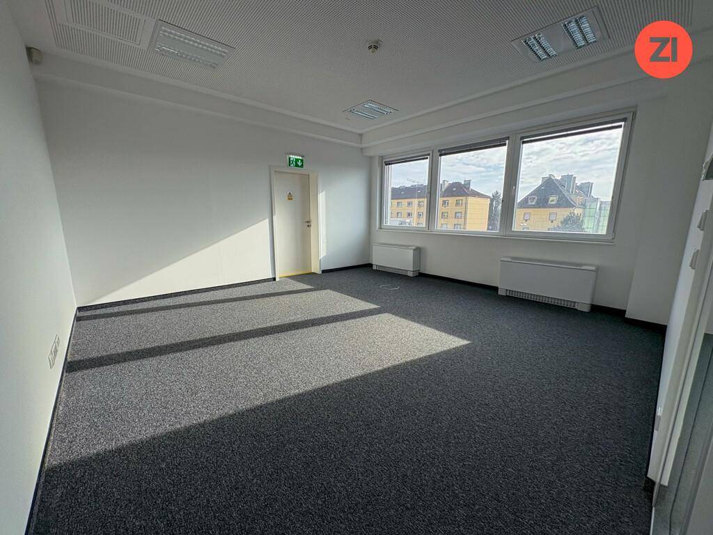 Leeres Büro mit Teppichboden, großen Fenstern und viel Sonnenlicht, das den Raum erhellt.